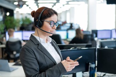 Kulaklık takmış güzel beyaz kadın uzay ofisinde dururken elinde bir cep telefonu tutuyor. Dost canlısı bayan yardımcı operatör iş yerindeki akıllı telefonun ekranına göz atıyor.