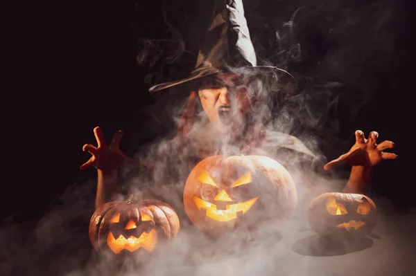 Una bruja siniestra con sombrero evoca sobre una linterna de gato. Personajes tradicionales de Halloween. Niebla mística se arrastra sobre calabazas con caras terribles talladas. Una bruja malvada lanza un hechizo. — Foto de Stock