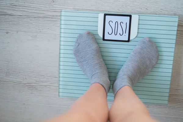 The fat woman is weighed. A top view of female feet in gray socks stands on an electronic scale. SOS inscription on the display of the floor scale. — Stock Photo, Image