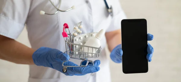 Uma médica sem rosto em um casaco médico com um estetoscópio segura um carrinho de compras mini cheio de diferentes medicamentos e um smartphone com uma tela em branco. Aplicação móvel para a compra de — Fotografia de Stock