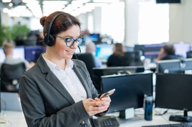 Kulaklık takmış güzel beyaz kadın uzay ofisinde dururken elinde bir cep telefonu tutuyor. Dost canlısı bayan yardımcı operatör iş yerindeki akıllı telefonun ekranına göz atıyor.