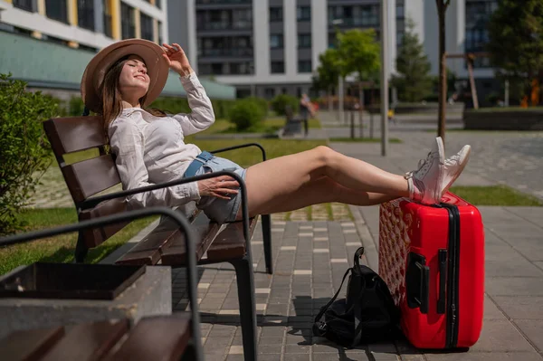 Mooie blanke vrouw met een hoed en korte broek zit op een bank haar benen te strekken op een rode koffer en te wachten op een taxi. Meisje wachtend op vertrek op een zomerreis. — Stockfoto
