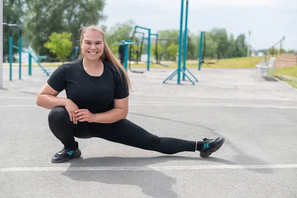 Fettleibige junge Kaukasierin beim Seitensprung. Dicke schöne lächelnde Mädchen in einem schwarzen Trainingsanzug ist in Fitness im Freien beschäftigt. Aufwärmen vor dem Training. — Stockfoto