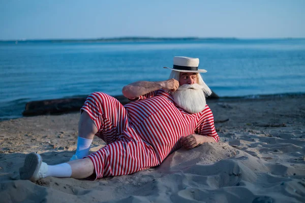 Um homem idoso de fato de banho retrô listrado toma banho de sol na praia. Um velho barbudo de cabelos grisalhos num chapéu jaz na areia junto ao mar.. — Fotografia de Stock