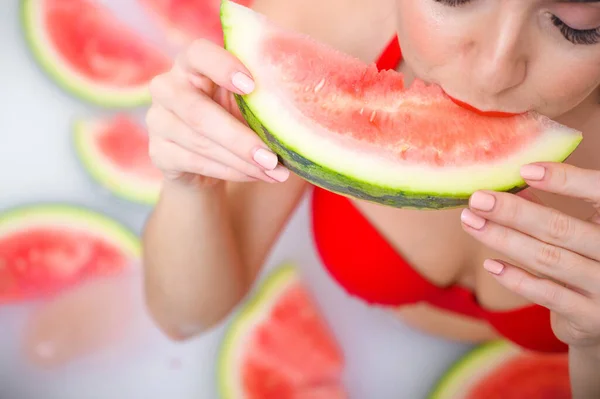 Portrait of a woman in a red swimsuit eating a watermelon and smirking. Redhead girl with red lipstick takes a bath with milk and fruit to rejuvenate her skin. Body care. — Stock Photo, Image