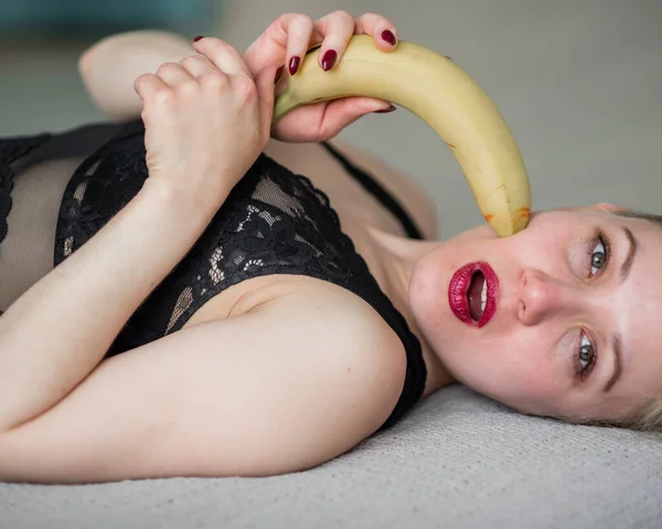 De blonde in zwart kanten ondergoed eet erotisch een banaan. Aantrekkelijke vrouw met sensuele rode lippen zuigt en likt seksueel een banaan. Oraal genot. — Stockfoto