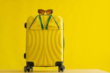 Tekerlekli sarı bavul, kaplan güneş gözlüğü ve stüdyoda sarı arka planda mini bir bikini. Plaj tatiline hazır olun. Uzayı kopyala İnsan yok. Denizde yaz tatili.