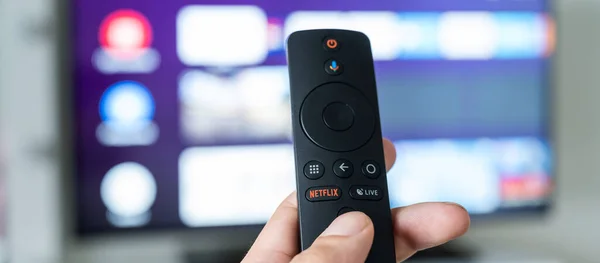 A man holds a remote control in his hand with the netflix logo on the background of a blurred television. Portal with unlimited access to film and TV shows. — Stock Photo, Image