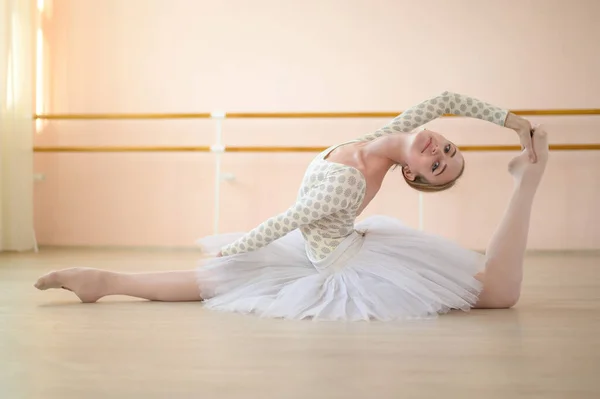 Balerina indah dalam tubuh dan tutu putih adalah pelatihan di kelas tari. Muda penari fleksibel berpose dalam sepatu runcing duduk di lantai. — Stok Foto