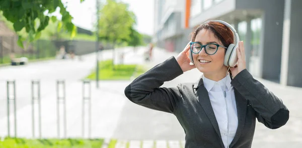 Retrato de una mujer sonriente en un traje de trabajo escuchando música en auriculares al aire libre. Empleada de oficina toma las manos en los auriculares disfrutando de la canción y bailando en la calle. —  Fotos de Stock