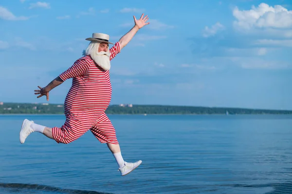 En äldre man i randig retro baddräkt springer och hoppar längs stranden. En glad gråhårig gammal man med skägg i hatt vilar vid havet.. — Stockfoto