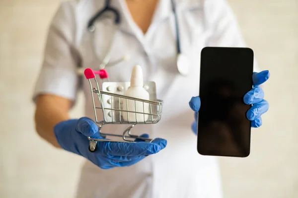 Egy arctalan női orvos egy sztetoszkóppal ellátott orvosi köpenyben tart egy mini bevásárlókocsit tele különböző gyógyszerekkel és egy okostelefont üres képernyővel. Mobil alkalmazás vásárlására — Stock Fotó