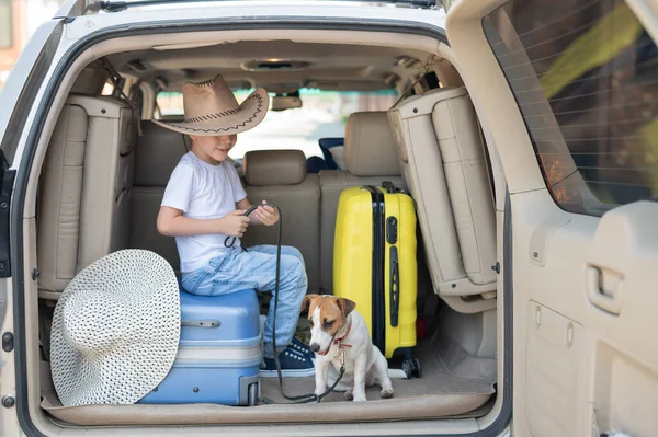 Glücklicher Junge mit Cowboyhut und Welpe Jack Russell Terrier reisen mit dem Auto. Ein Kind und ein lustiger kleiner Hund sitzen im Kofferraum und sind bereit für die Sommerferien. Unabhängiges Reisen. Beste Freunde. — Stockfoto