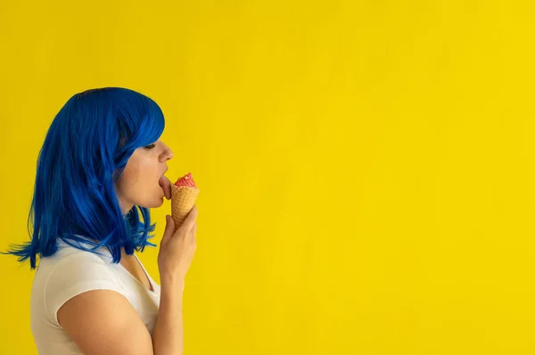 Une belle femme aux cheveux bleus se dresse de profil et lèche une corne de glace à la framboise sur fond jaune. La fille à la perruque aime manger de la gelée dans une tasse à gaufres. dessert de refroidissement d'été. — Photo