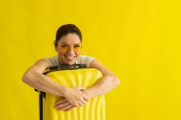 一个戴着太阳镜，雪白的笑容完美的女人抱着一个黄色背景的手提箱。兴奋地等待暑假旅行的女孩依偎在她的包里. — 图库照片