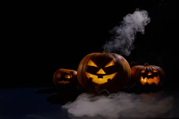 Three creepy halloween steaming pumpkins with a carved luminous smirk on a black background. A handmade jack-o-lantern head with a candle inside in the dark among the fog. Trick or treat. Postcard. — Stock Photo, Image