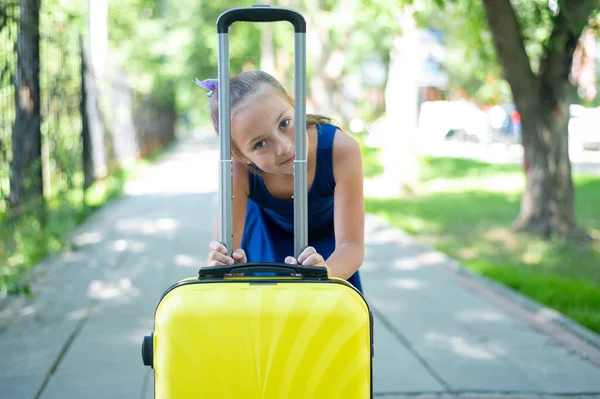 Een tevreden meisje in een blauwe jurk staat alleen op straat met een gele koffer en wacht op haar ouders. Een kind gaat op zomervakantie.. — Stockfoto