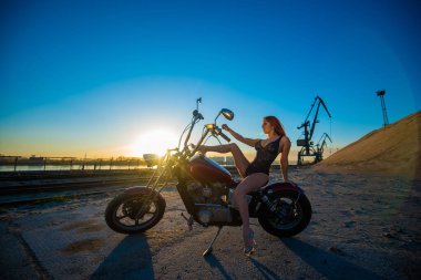 Yüksek topuklu, erotik iç çamaşırlı genç, kızıl saçlı bir kadın motosiklette oturuyor. Bisikletli seksi bir kızın silueti. Sanayi manzarası.