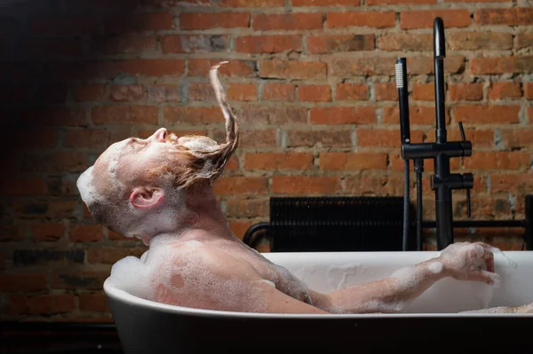 Close-up portrait of funny red-bearded bald man takes a bath with foam on the background of a brick wall. Cheerful naughty guy at spa treatments. A parody of glamorous girls. — Stock Photo, Image