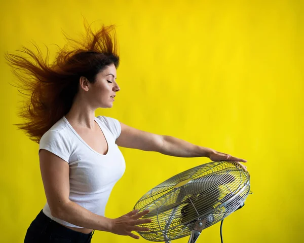 Una hermosa mujer pelirroja se enfría de pie sobre un gran ventilador eléctrico sobre un fondo amarillo. Chica con el pelo en desarrollo en el viento. Dispositivo para enfriar el aire. — Foto de Stock