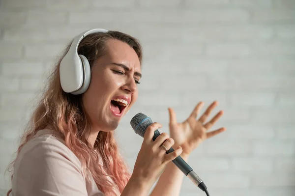 Közelkép egy göndör hajú kaukázusi nőről, aki mikrofonba énekel. Gyönyörű érzelmi lány fehér fülhallgatóval énekel egy dalt otthon karaoke és aktívan gesztusok ellen téglafal. — Stock Fotó