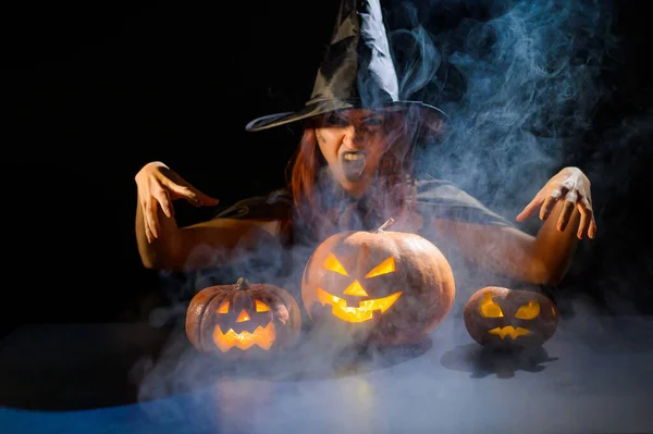 Una bruja siniestra con sombrero evoca sobre una linterna de gato. Personajes tradicionales de Halloween. Niebla mística se arrastra sobre calabazas con caras terribles talladas. Mujer disfrazada de carnaval. — Foto de Stock