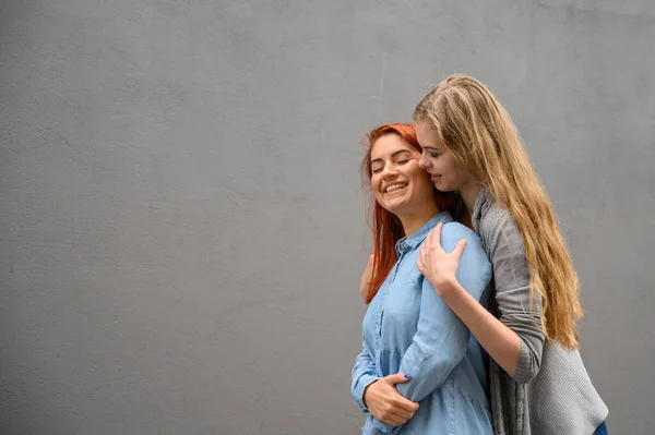 Twee blije vriendinnen knuffelen op de achtergrond van een grijze muur. Zachte knuffels van een lesbisch echtpaar. LGBT huwelijk van hetzelfde geslacht. — Stockfoto