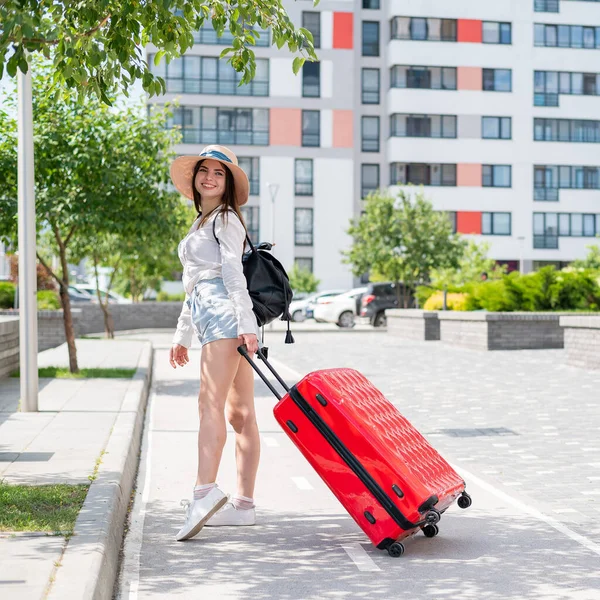 Schöne junge kaukasische Frau mit Sommermütze posiert mit einem roten Koffer im Freien. Glücklich lächelndes Mädchen geht mit einer großen Gepäcktasche in den Urlaub. Charmante Dame in kurzen Hosen an einem heißen, sonnigen Tag. — Stockfoto