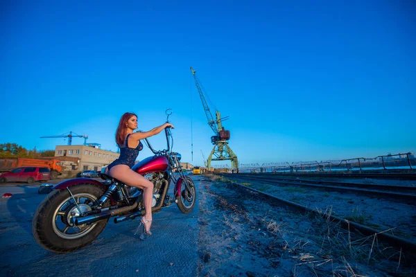 Sexy jovem mulher ruiva em roupa interior erótica senta-se em uma motocicleta vermelha. Menina sedutora dirige uma bicicleta em um fundo industrial . — Fotografia de Stock