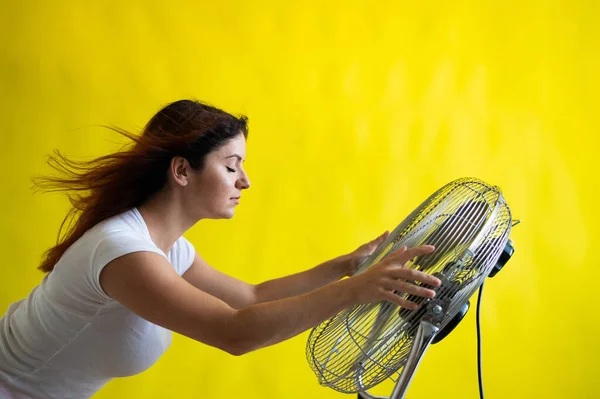 Una hermosa mujer pelirroja se enfría de pie sobre un gran ventilador eléctrico sobre un fondo amarillo. Chica con el pelo en desarrollo en el viento. Dispositivo para enfriar el aire. — Foto de Stock