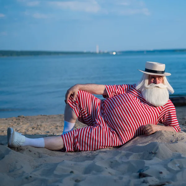 Un uomo anziano in costume da bagno retrò a righe prende il sole sulla spiaggia. Un vecchio uomo barbuto dai capelli grigi con un cappello giace sulla sabbia vicino al mare. — Foto Stock