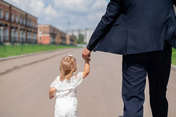 Вид сзади на человека в черном костюме прогуливается со своей маленькой дочкой на улице. Маленькая девочка в белом платье держит своих отцов за руку. Отцовская любовь — стоковое фото