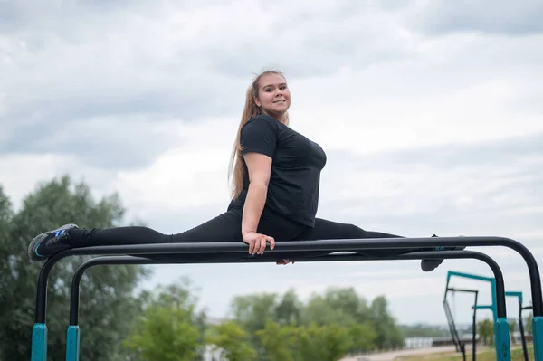 Eine schöne lächelnde, übergewichtige junge Frau streckt sich auf unebenen Stufenbarren im Freien. Dicke Turnerinnen beim Fitnesstraining auf dem Sportplatz. Sport draußen an einem warmen Sommertag. — Stockfoto