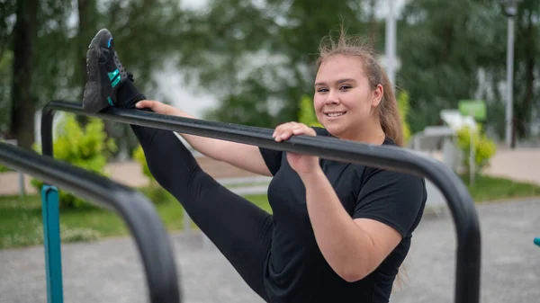 Tlustá dívka se zabývá protáhnutím na nerovné bary na hřišti. Krásná mladá usměvavá žena dělá sport pro hubnutí venku. — Stock fotografie