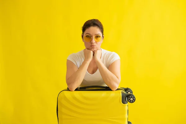 戴太阳镜的不快乐女人抱着一个黄色背景的手提箱.一个心烦意乱的女孩在飞机上错过了她的航班.取消了所有与头孢病毒有关的航班。没有暑假. — 图库照片