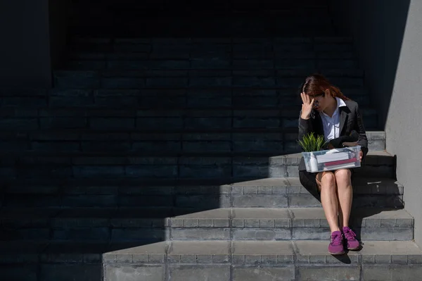 Mujer deprimida vestida con máscara médica es despedida y está sentada en las escaleras con una caja de pertenencias personales. Trabajadora de oficina con traje y zapatillas al aire libre. El desempleo en la crisis económica —  Fotos de Stock