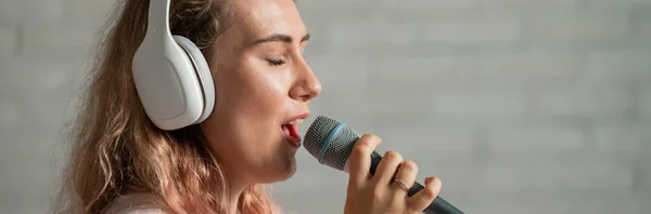 Portret kaukaskiej kobiety z kręconymi włosami śpiewającej do mikrofonu. Piękna zmysłowa dziewczyna w białych słuchawkach śpiewa piosenkę w domu karaoke. — Zdjęcie stockowe