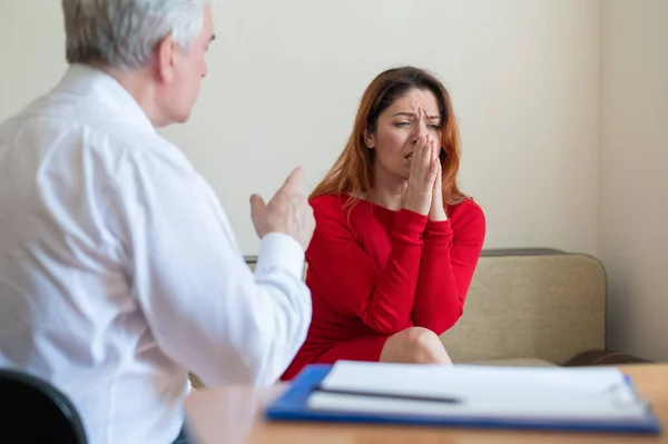 A frustrated crying woman holds her palms over face at a session with a male psychologist. Mature psychotherapist talking to a female patient with depression and neurosis. Mental health treatment.