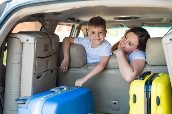 Mulher europeia feliz com seu filho pequeno olhar para trás na bagageira de um carro com malas. Férias em família. Viajar de carro com uma criança. Um rapaz com a mãe no banco de trás de um jipe . — Fotografia de Stock