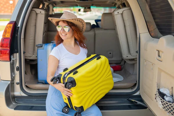 Glückliche rothaarige Frau mit Cowboyhut und Sonnenbrille auf einer Autofahrt durch das Land. Mädchen im Kofferraum mit gelbem Koffer, bereit für den Sommerurlaub. Unabhängiges Reisen. — Stockfoto