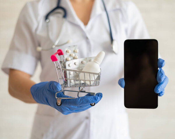 A faceless female doctor in a medical coat with a stethoscope holds a mini shopping trolley full of different medicines and a smartphone with a blank screen. Mobile application for the purchase of