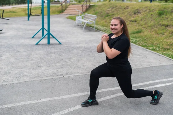 Uma linda garota gorda em um traje preto está envolvida na aptidão no campo esportivo. Jovem mulher lunges ao ar livre em um dia quente de verão. Estilo de vida saudável e perda de peso. — Fotografia de Stock