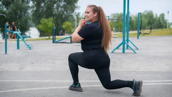 一个穿着黑色运动服的漂亮的胖女孩正在运动场上健身。在一个温暖的夏日,年轻的女人在户外吃午饭.健康的生活方式和减肥. — 图库照片