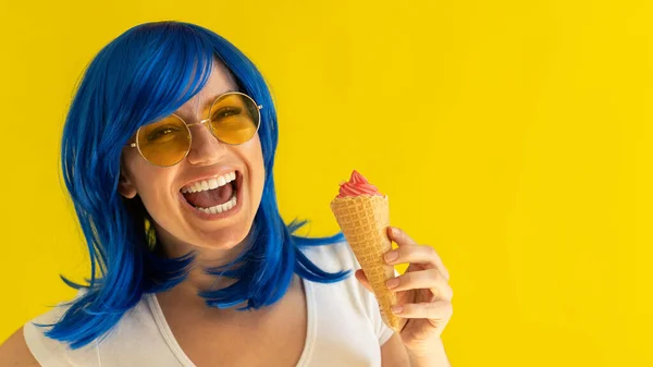 Porträtt av en kvinna med blått hår i solglasögon håller en våffla kon med glass på en gul bakgrund. En vacker vit flicka med ett perfekt leende njuter av en sommar kylande dessert. — Stockfoto