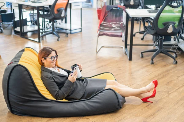 Business woman resting in a cozy bag chair and enjoys mobile. Female Manager at a break in a modern office. Caring for the convenience of employees. Beanbag in the workplace.