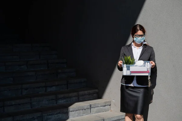 unemployed woman in a protective mask is standing with a box of personal belongings at the stairs. Business lady fired. The global economic crisis. Reduction of work due to the companys bankruptcy.