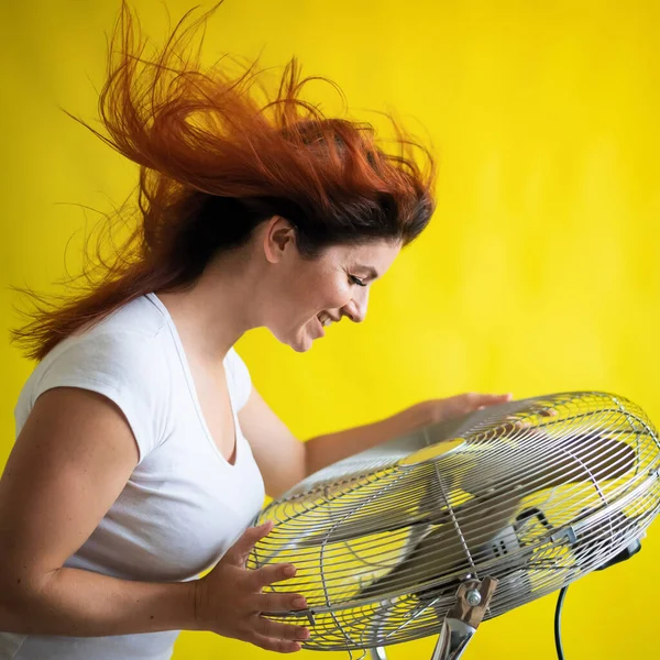 Una hermosa mujer pelirroja se enfría de pie sobre un gran ventilador eléctrico sobre un fondo amarillo. Chica con el pelo en desarrollo en el viento. Dispositivo para enfriar el aire. — Foto de Stock