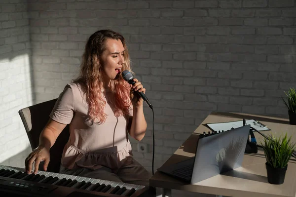 Een jonge Europese vrouw zingt in een microfoon en begeleidt op een elektronische piano. Het meisje maakt een videoblog. Zang op afstand. Een zanglerares geeft online les. — Stockfoto