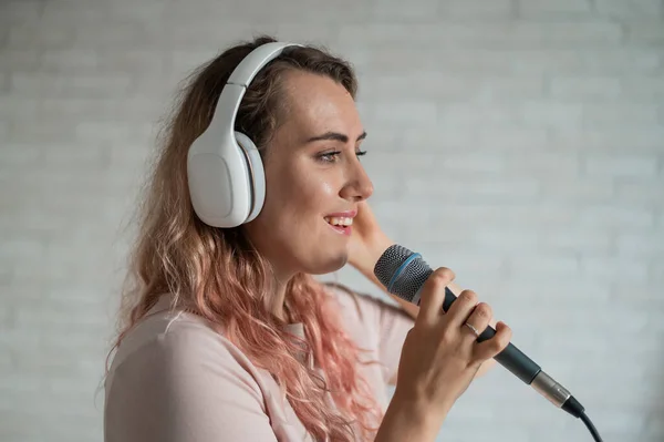 Großaufnahme Porträt einer kaukasischen Frau mit lockigem Haar, die in ein Mikrofon singt. Schöne sinnliche Mädchen in weißen Kopfhörern singt ein Lied in Karaoke zu Hause. — Stockfoto