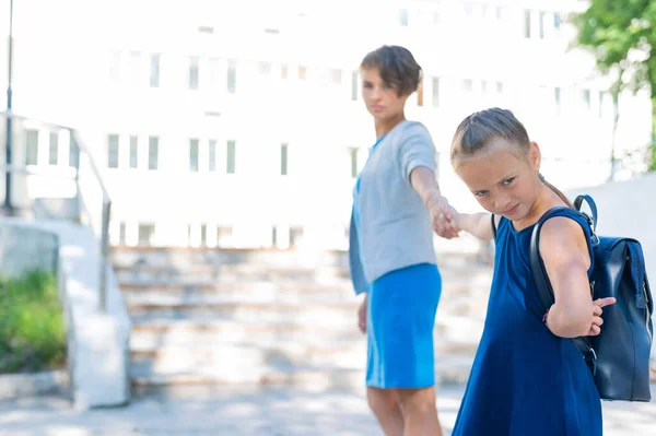 Malá nešťastná holka s batohem tahá matku za ruku a nechce jít do školy. Žena vede vzdorující dceru do školy. Konflikt generací. — Stock fotografie